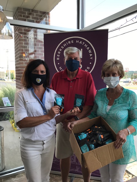 Dale Meller and two other females, one on his right is blonde wearing a teal dress top clutching a box full of small pouches. The brunette female on his left is wearing white and holding two of the pouches to the camera. All three are wearing masks